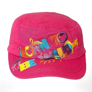 Puede incluir: Una gorra de estilo militar de color rosa brillante con texto bordado multicolor que dice BORN TO BE WILD. El diseño incluye figuras de animales vibrantes y cosidas, como una jirafa y un caballo, integradas en las letras del panel frontal.