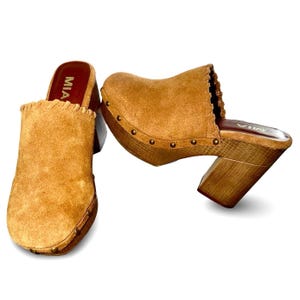 Pode incluir: Um par de tamancos de camurça castanhos com saltos de madeira. Os tamancos têm bico arredondado e uma borda ondulada ao redor da abertura. Os saltos têm aproximadamente 10 cm de altura e são cravejados com pequenos detalhes metálicos redondos.