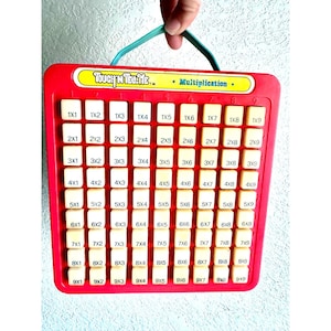 May include: A red multiplication learning tool with a green handle. The board has a grid of white tiles with multiplication problems. The text "TOUCH 'N TELL ME" is at the top.