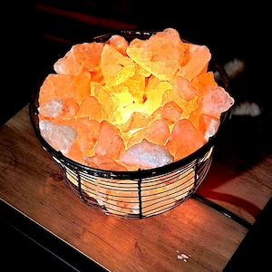 May include: A round, black metal basket filled with glowing, orange Himalayan salt chunks. The salt crystals are illuminated from within, creating a warm, ambient glow. The basket sits on a wooden surface.