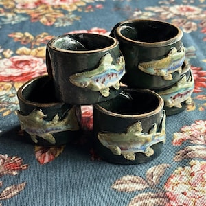 May include: A set of four cylindrical ceramic napkin rings with a dark, glossy black glaze. Each ring features a raised, three-dimensional relief of a fish with iridescent, multi-colored scales in shades of blue, green, and tan.