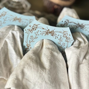May include: Several light blue wooden decorative pieces with a floral design in brown are arranged on top of white fabric. The pieces have a curved shape with a cutout in the center.