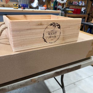 May include: A rectangular light-colored wooden crate featuring a black laser-engraved design on the front. The design includes a floral wreath surrounding the text "the WEST family" and the phrase "EVERY FAMILY HAS A STORY". It has rope handles.