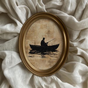 May include: An oval-shaped wall art piece featuring a black silhouette of a person fishing from a small boat. The artwork is set against a textured, light tan background and is enclosed within a smooth, metallic gold-toned oval frame.