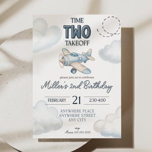 May include: A light beige invitation card with a watercolor illustration of a biplane and clouds. The text reads "TIME TWO TAKEOFF" and "Miller's 2nd Birthday" with event details. The card is on a white surface with shadows.