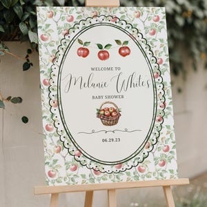May include: A wooden easel displays a baby shower sign. The sign features a white oval with the text "Welcome to Melanie White's BABY SHOWER" and a basket of apples. The background is a pattern of apples.