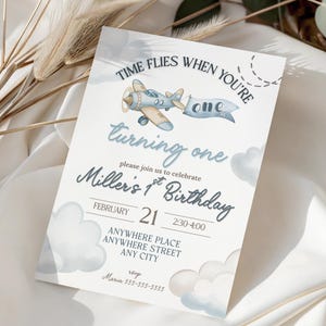 May include: A light blue and beige airplane illustration on a white invitation card. The card reads "Time flies when you're turning one" and "Miller's 1st Birthday" with event details. Decorative clouds and text are in blue.