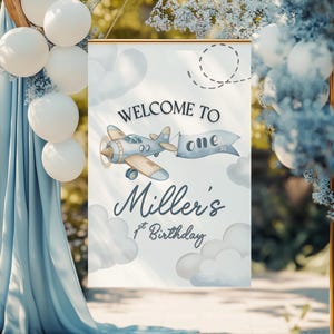 May include: A rectangular sign with a gold frame features the text "WELCOME TO ONE" and "Miller's 1st Birthday". An illustration of a vintage airplane with a banner is included. The background has clouds and a dotted line. White balloons and blue fabric are in the background.