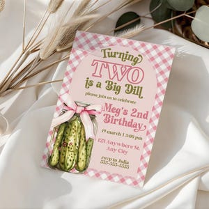 May include: A pink and white gingham birthday invitation with the text "Turning TWO is a Big Dill." The invitation features a jar of pickles and a pink bow. The invitation includes the birthday details.