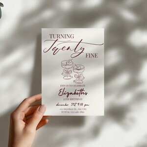 Puede incluir: Una tarjeta de invitaci&oacute;n blanca con texto e ilustraciones en color burdeos. La tarjeta dice "Turning Twenty Fine" e incluye detalles para una celebraci&oacute;n de 25 cumplea&ntilde;os, con copas de c&oacute;ctel. La tarjeta est&aacute; sujeta por una mano.