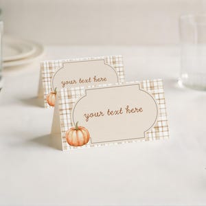 May include: Two folded place cards with a pumpkin illustration. The cards have a beige background with a plaid pattern and a decorative frame. The text "your text here" is printed in brown. The cards are on a white surface.