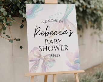 May include: A baby shower sign on a wooden easel. The sign reads "Welcome to Rebecca's Baby Shower 08.14.25" in elegant script. The design includes watercolor elements of a mermaid tail and a bow in pastel colors.