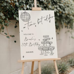 以下が含まれることがあります： 木製のイーゼルに立てられたクリーム色の看板。黒い文字とイラストが描かれています。「a tiny bit older」と書かれており、Brookeの30歳の誕生日にゲストを歓迎します。01.30.2025。ディスコボールとシャンパンタワーが描かれています。