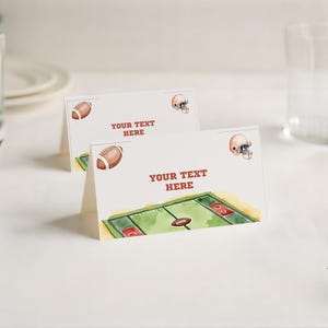 Op de afbeelding: Twee gevouwen naambordjes met een voetbalthema. Elke kaart heeft een aquarel illustratie van een voetbal, een voetbalhelm en een voetbalveld. De tekst "YOUR TEXT HERE" is rood gedrukt. De kaarten liggen op een wit oppervlak.