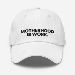 May include: A white baseball cap featuring the black embroidered text MOTHERHOOD IS WORK. on the front panel. The hat has a structured crown, a curved brim, and a button on top, providing a clean and simple design for everyday headwear.