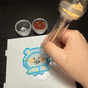 Puede incluir: Una herramienta de plástico transparente se utiliza para colocar pequeñas cuentas de color claro en una placa de clavijas blanca para crear un diseño de oso azul y blanco. Dos pequeños recipientes transparentes contienen cuentas marrones y claras.