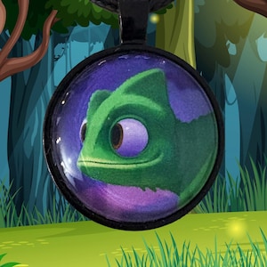 May include: A round pendant necklace featuring a green chameleon with purple eyes against a purple and blue background. The pendant is suspended from a black cord necklace. The background is a cartoon forest scene with trees, grass, and mushrooms.