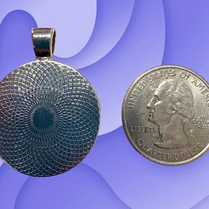 Puede incluir: Colgante plateado con dise&ntilde;o circular, junto a una moneda de veinticinco centavos. El colgante tiene un lazo para una cadena. La moneda muestra un perfil de George Washington.