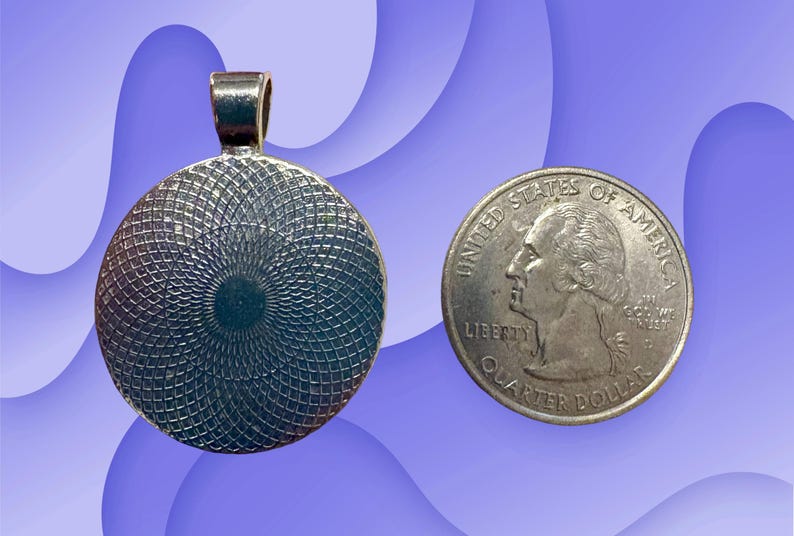 Puede incluir: Colgante plateado con dise&ntilde;o circular, junto a una moneda de veinticinco centavos. El colgante tiene un lazo para una cadena. La moneda muestra un perfil de George Washington.