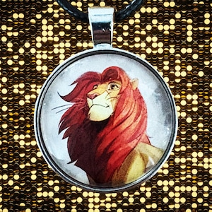 May include: Round pendant necklace featuring a lion's head with a red mane and a silver-colored metal frame. The pendant hangs from a black cord. The lion's face is detailed with a light brown and tan color scheme.