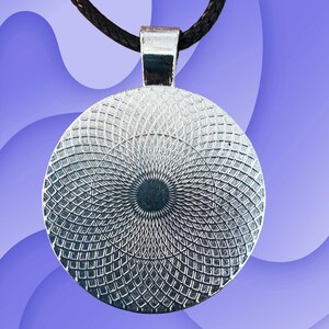 Puede incluir: Primer plano de un colgante plateado con un patr&oacute;n geom&eacute;trico. El colgante redondo est&aacute; unido a un collar de cord&oacute;n negro. El fondo es un degradado de morado y azul.