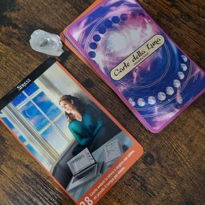 Könnte beinhalten: Ein Kartendeck "Carte della Luna" Tarotkarten mit einem lila und orangefarbenen Design, neben einer Karte mit dem Wort "STRESS" und einer Frau, die aus einem Fenster schaut. Ein klarer Kristall liegt in der Nähe. Die Karten liegen auf einer Holzoberfläche.