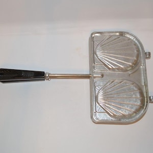 May include: A silver-colored metal shell-shaped cookie iron with two distinct molds for baking. The cooking tool features a long, thin metal rod connected to a black, faceted plastic handle, designed for creating two scallop-shaped treats.