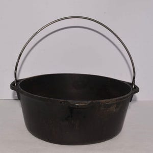 May include: A round, black cast iron pot featuring a wide, open top and a sturdy, arched metal bail handle attached to two side lugs. The exterior surface shows a dark, weathered patina with subtle variations in texture and color throughout.