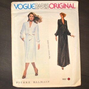 May include: A Vogue Paris Original sewing pattern envelope for Pierre Balmain designs, featuring illustrations of a white knee-length dress and a black floor-length gown with long sleeves and tie-neck details, marked with the number 1857.