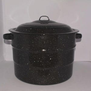 May include: A large, cylindrical black stockpot with a speckled white pattern throughout the exterior. The pot features two side handles, a matching lid with a small arched handle on top, and two horizontal ridges encircling the main body.