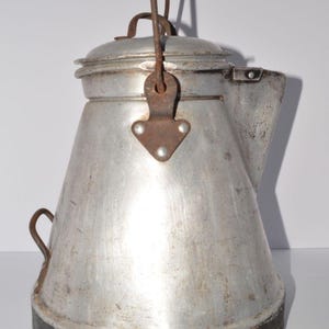 May include: A vintage aluminum coffee percolator with a tapered body, a dark base, and a rusted metal bail handle. The pot features a lid with a small loop handle, a side spout, and a small metal loop on the lower side, showing signs of wear.