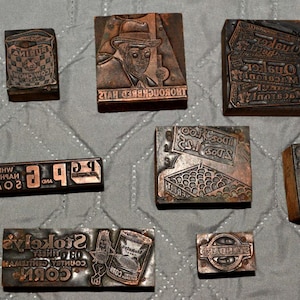 May include: A collection of vintage copper printing blocks with various designs and text. The blocks feature images of a man in a hat, a bag of goods, and text including "WHITE SOAP" and "STOKES' COUNTRY GENTLEMAN CORN".