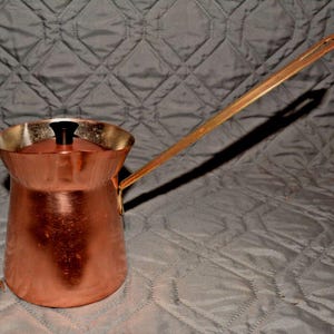 May include: A copper-colored Turkish coffee pot with a long, wooden handle. The pot has a rounded base, a flared rim, and a black lid. The handle extends from the side, offering a classic design for brewing coffee.