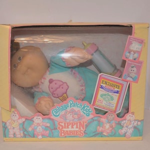 May include: A Cabbage Patch Kids Sippin Babies doll in its original yellow cardboard box. The doll wears a teal outfit with a white bib featuring a pink ice cream cone graphic. It holds a plastic baby bottle with a pink cap and base.