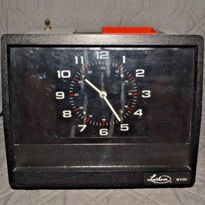 May include: A black Lathem 8100 clock with a rectangular face. The clock face has white numerals and hands, with red accents. A small red piece sits on top of the clock.