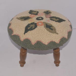 May include: A round footstool featuring a textured, cream-colored hooked rug top with a floral pattern in shades of dark green, olive, and soft pink. The stool is supported by four turned wooden legs with a smooth, medium-brown finish.