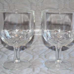 May include: Two clear, stemmed wine glasses. The bowl of each glass has a rounded shape with a textured pattern. The glasses sit on a round base and have a slender stem. The background is a light gray fabric.
