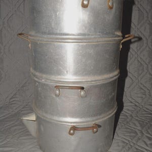 May include: A vintage aluminum coffee percolator with a three-tiered design. It features a lid with a black knob and side handles for easy handling. The percolator has a spout on the bottom tier.