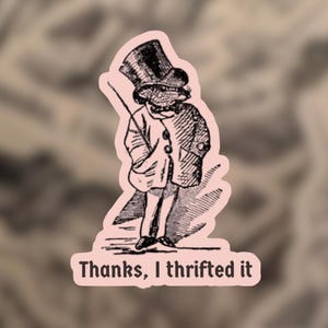 May include: A sticker featuring a vintage illustration of a frog in a top hat and coat, holding a cane. The text "Thanks, I thrifted it" is printed in a serif font at the bottom of the sticker. The sticker has a pink border.