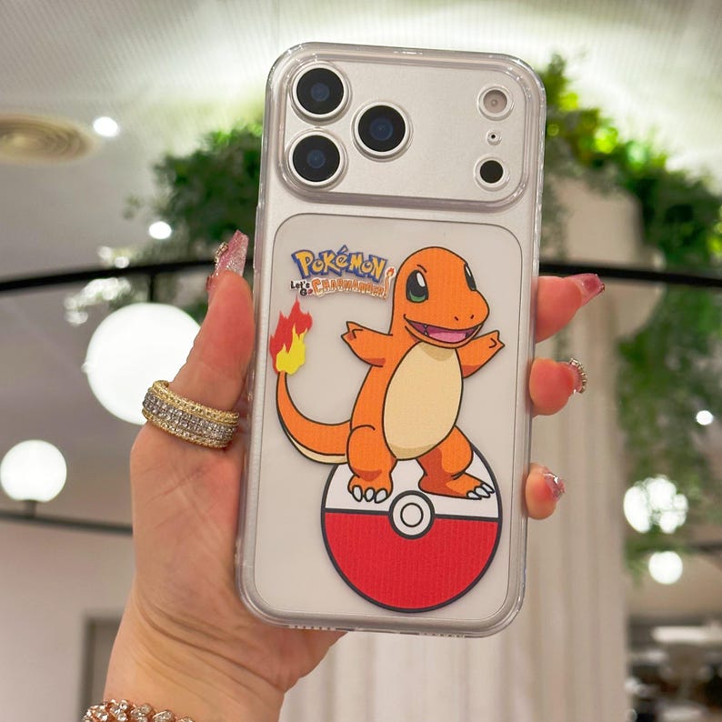 May include: Clear phone case featuring a cartoon Charmander character from Pok&eacute;mon. The orange Charmander is standing on a red and white Pok&eacute; Ball. The case has a clear border and the text "Pok&eacute;mon Let's Go!" is visible.