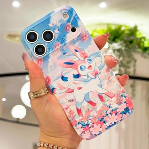 May include: A smartphone case with a blue and white design, featuring a cartoon Sylveon character with pink details. The case has a floral and sky background and a glossy finish.