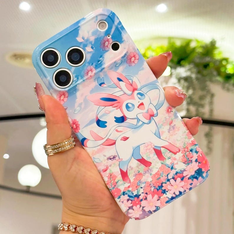 May include: A smartphone case with a blue and white design, featuring a cartoon Sylveon character with pink details. The case has a floral and sky background and a glossy finish.