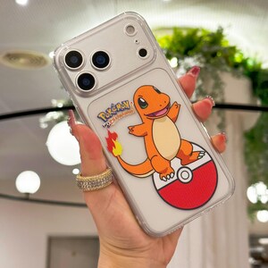 May include: Clear phone case featuring a Charmander design from Pok&eacute;mon. The orange and cream-colored character is standing on a red and white Pok&eacute; Ball. The case has a clear border and a silver phone back.