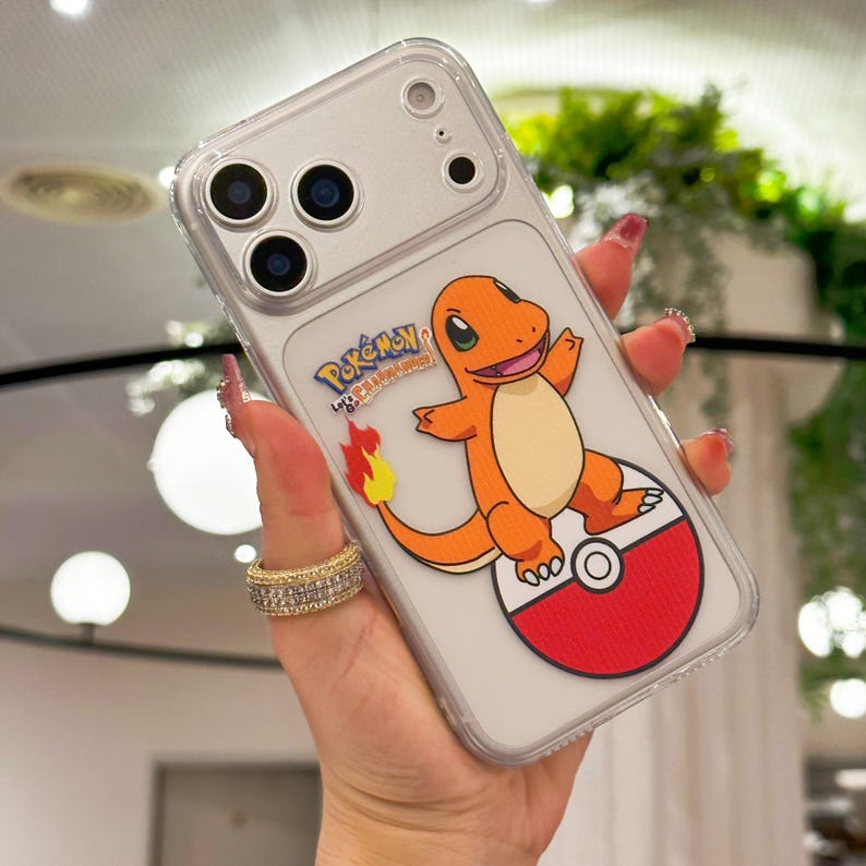 May include: Clear phone case featuring a Charmander design from Pok&eacute;mon. The orange and cream-colored character is standing on a red and white Pok&eacute; Ball. The case has a clear border and a silver phone back.