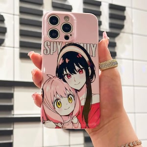 May include: Pink phone case featuring the text "SPY FAMILY" and an anime illustration of two characters. The case has a glossy finish and a camera cutout. The design includes a character with black hair and another with pink hair.