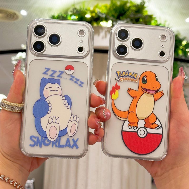 May include: Two clear phone cases with cartoon designs. One features a sleeping blue Snorlax with "Zzz" text, and the other showcases an orange Charmander on a red and white Pok&eacute; Ball. Both cases have a clear border.