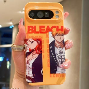 Op de afbeelding: Oranje telefoonhoesje met het woord "BLEACH" in grote oranje letters. De hoes heeft een afbeelding van twee anime-personages. De hoes is ontworpen voor een telefoon met een camera en flits.