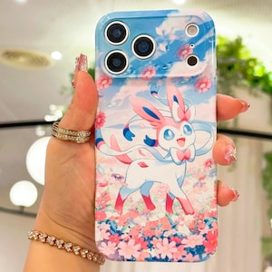 May include: A smartphone case with a Sylveon cartoon design. The case has a blue sky and flower background with pink and white accents. The case has a camera cutout. The case is designed for a smartphone.