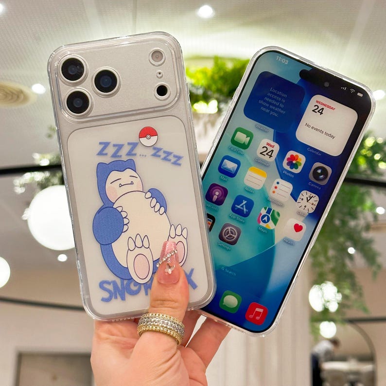 May include: Clear phone case featuring a blue and white cartoon Snorlax design with the text "SNOX" and "ZZZ". The phone case is held next to a phone displaying the date and time.