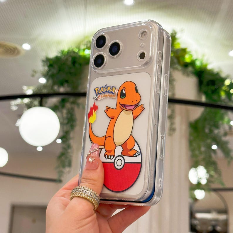 May include: Clear phone case featuring a cartoon Charmander character from Pok&eacute;mon. The orange and cream-colored Charmander is standing on a red and white Pok&eacute;ball. The case has a clear back and sides, with a silver camera module.